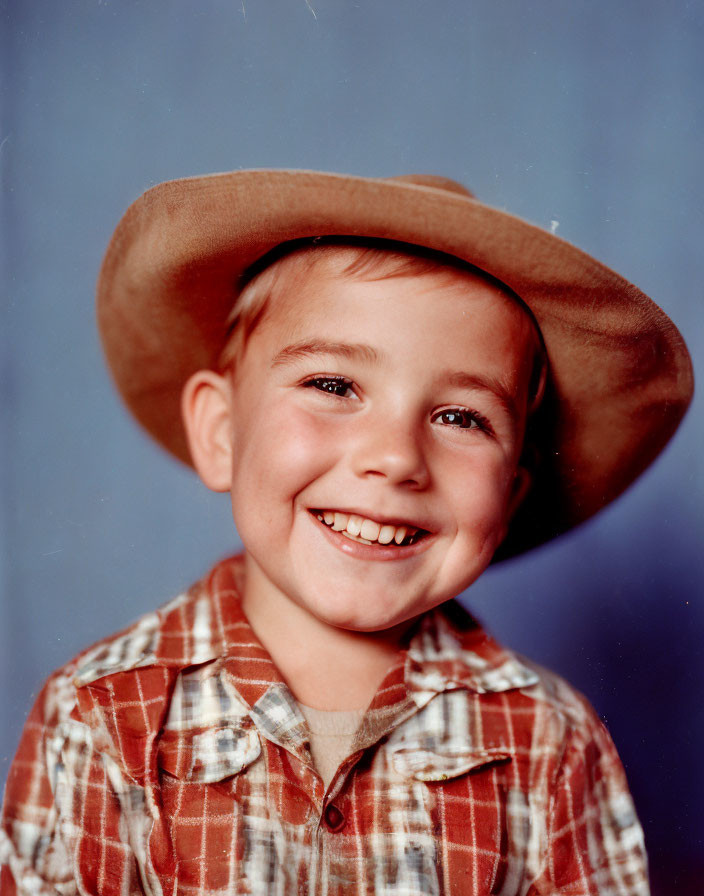 Young boy in cowboy hat and checkered shirt smiling on blue background