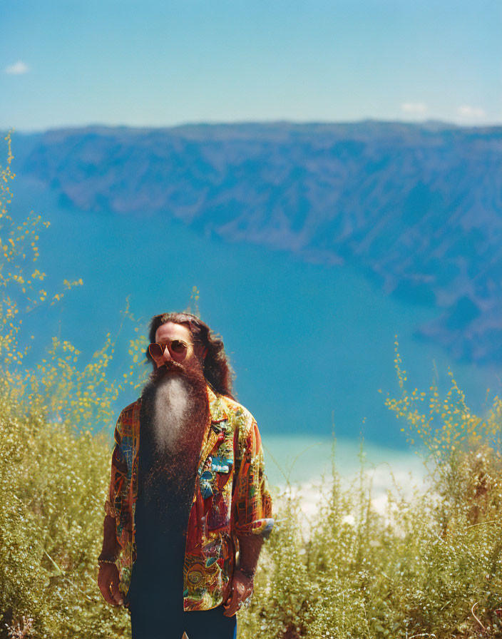 Bearded man in sunglasses by scenic lake and mountains with wildflowers