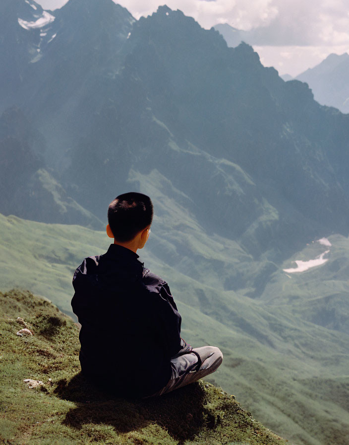 Person sitting on grassy hill, viewing majestic mountain range