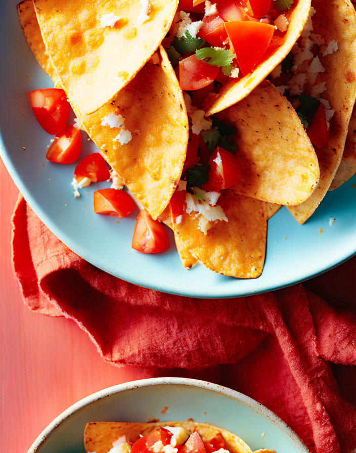 Three crispy tacos with cheese and diced tomatoes on a light blue plate, red napkin, coral background