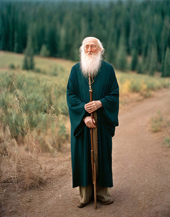 Elderly man with white beard in green robe on dirt road with staff