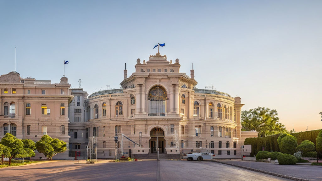 Neoclassical building with intricate facades and flags, surrounded by hedges and vehicles