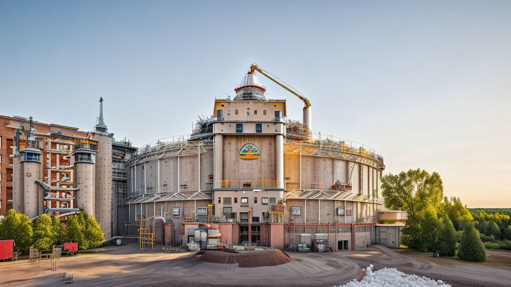 Industrial grain storage facility with silos, conveyor, and machinery in forested landscape at golden hour