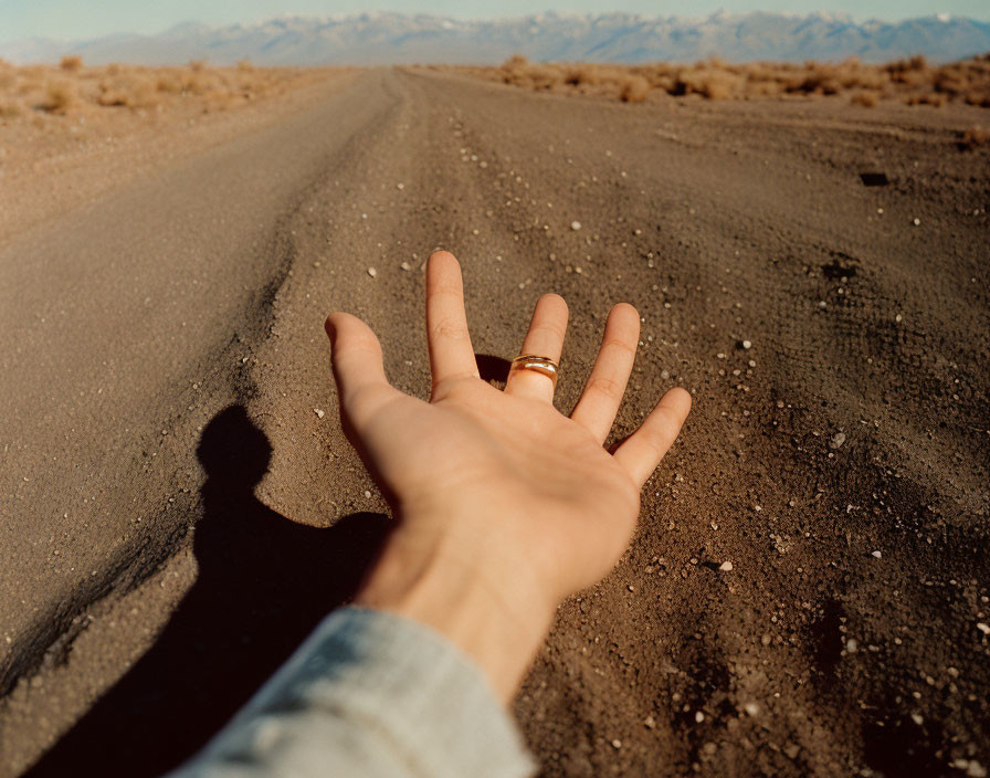 Hand with ring on finger on dirt road with shadow and mountains.