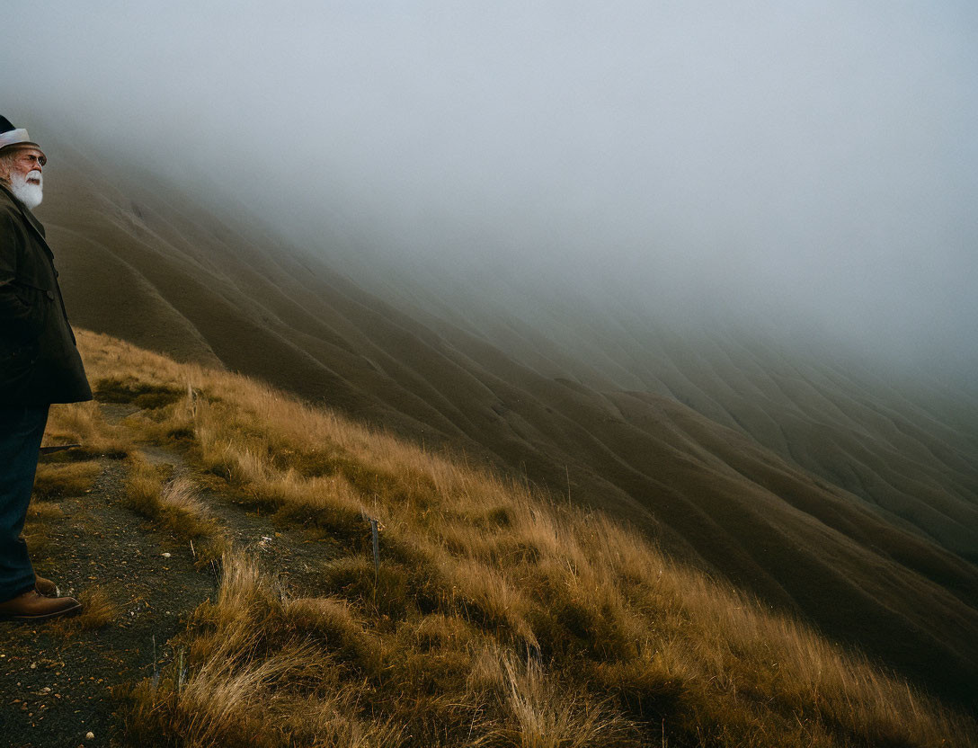 Bearded elderly man in jacket on grassy hill gazes into foggy landscape