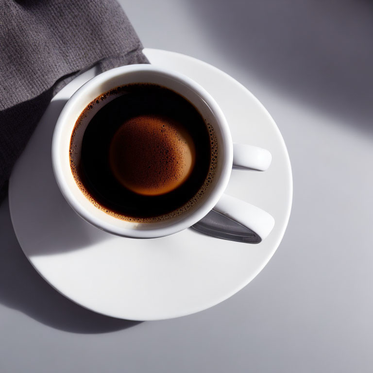 Top view of full black coffee cup with froth on white saucer and spoon