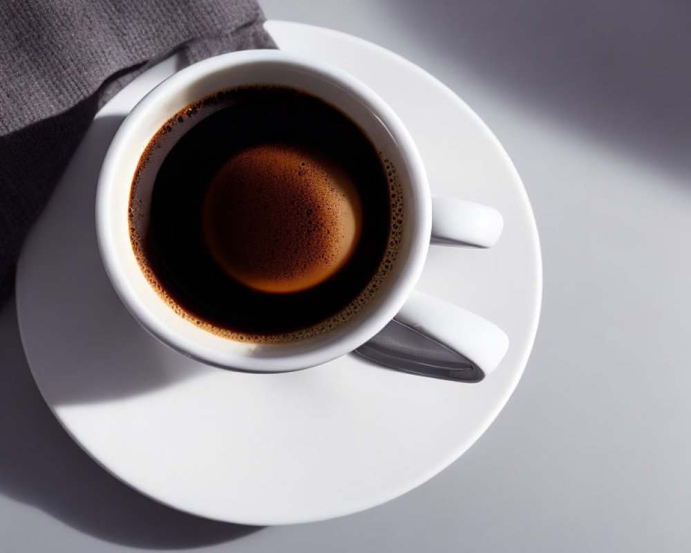 Top view of full black coffee cup with froth on white saucer and spoon