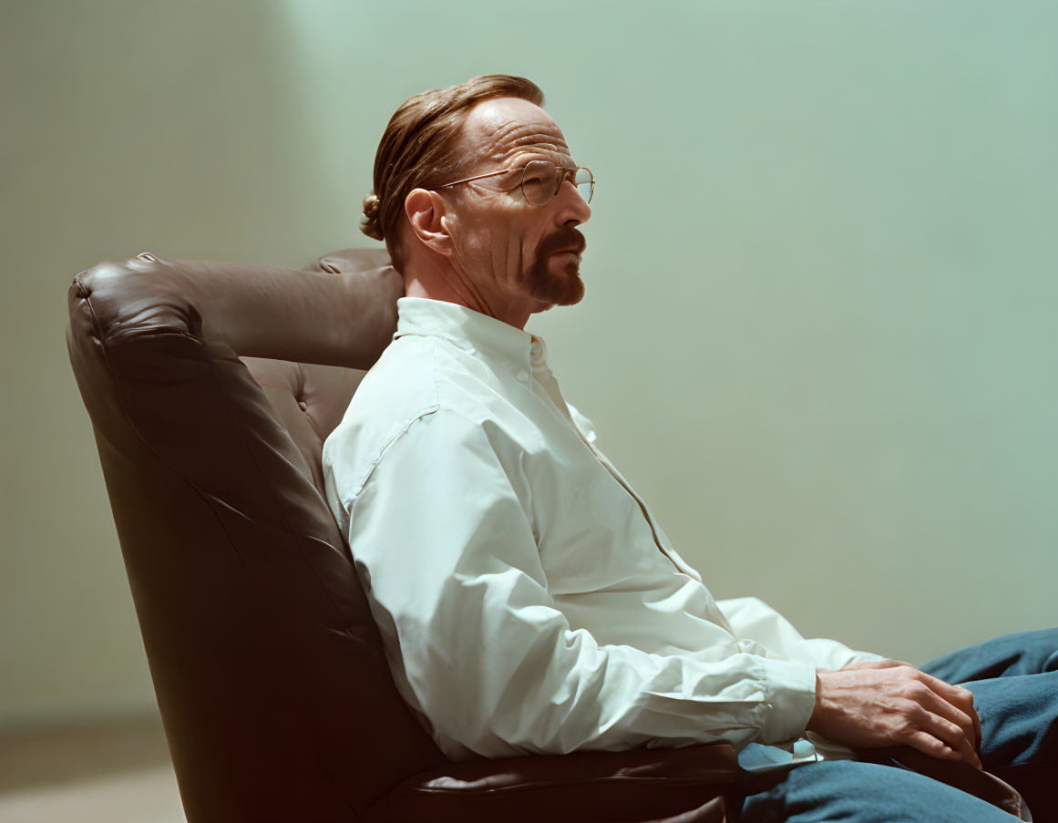 Man with glasses and mustache in white shirt and blue pants sitting in leather chair.