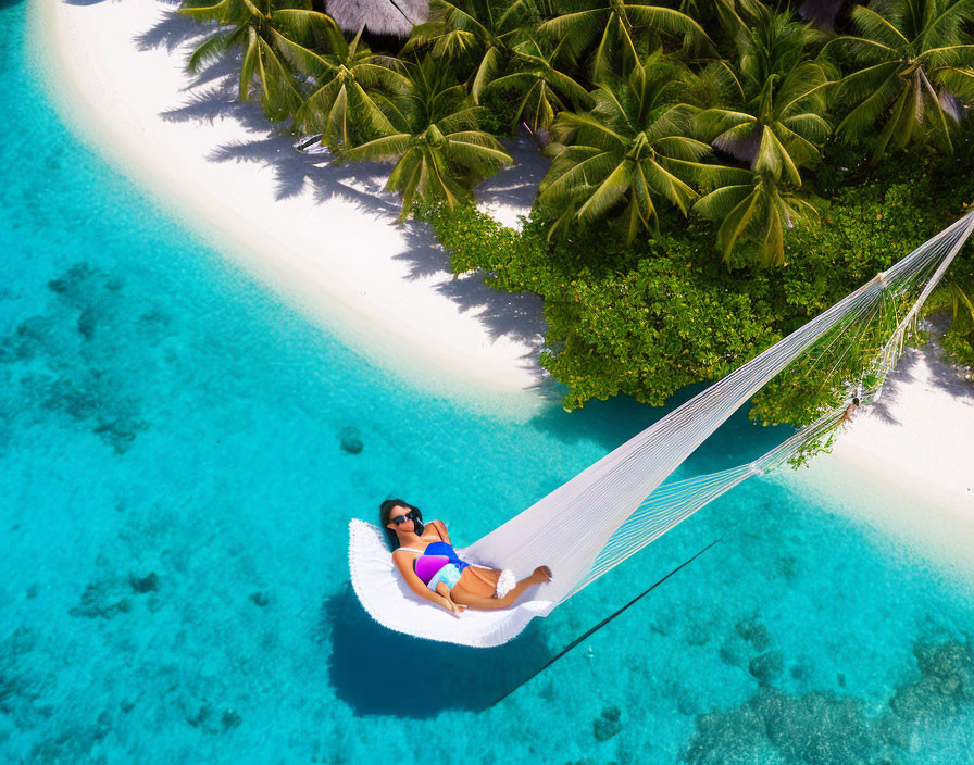 Person relaxing on hammock over clear blue waters near tropical island with lush palm trees.