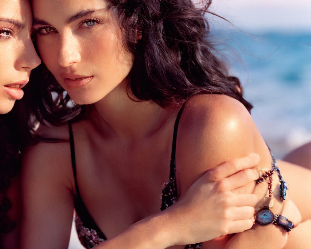 Two women in swimwear at the beach, one looking at the camera, the other gazing away