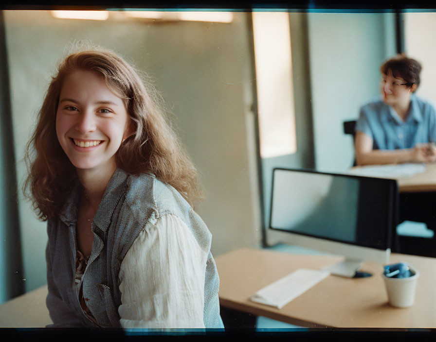 Smiling woman at desk with another person and monitor in background