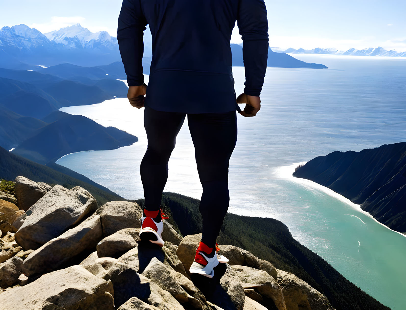 Person admires mountain summit view with fjord, snow-capped peaks, and blue sky