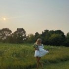 Young girl in dress running through grassy field at sunset with flowers and trees.