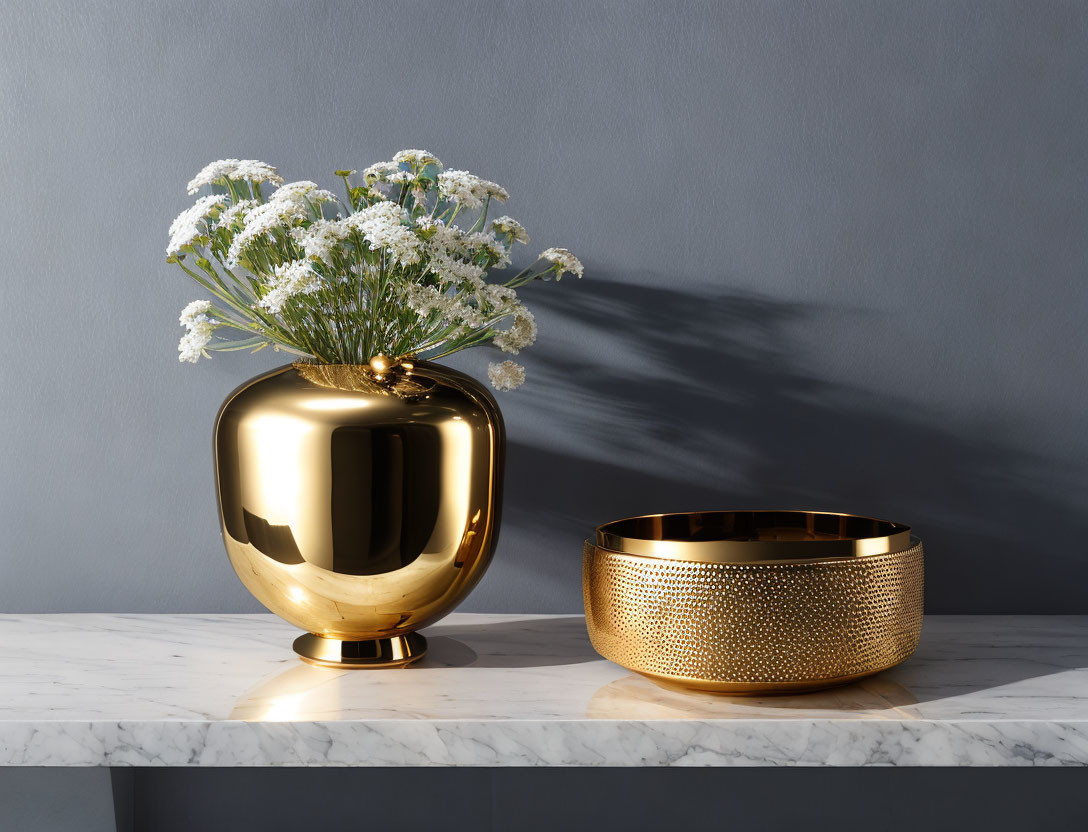 Golden vase and bowl with white flowers on marble countertop against grey wall