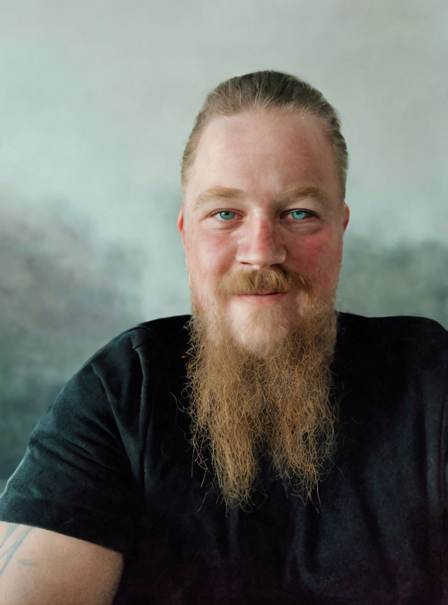 Smiling person with long beard, mustache, blue eyes, and tattoo on arm