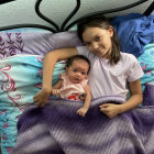 Two children in bed under purple blanket, one with sleep mask holding marshmallow, other smiling.