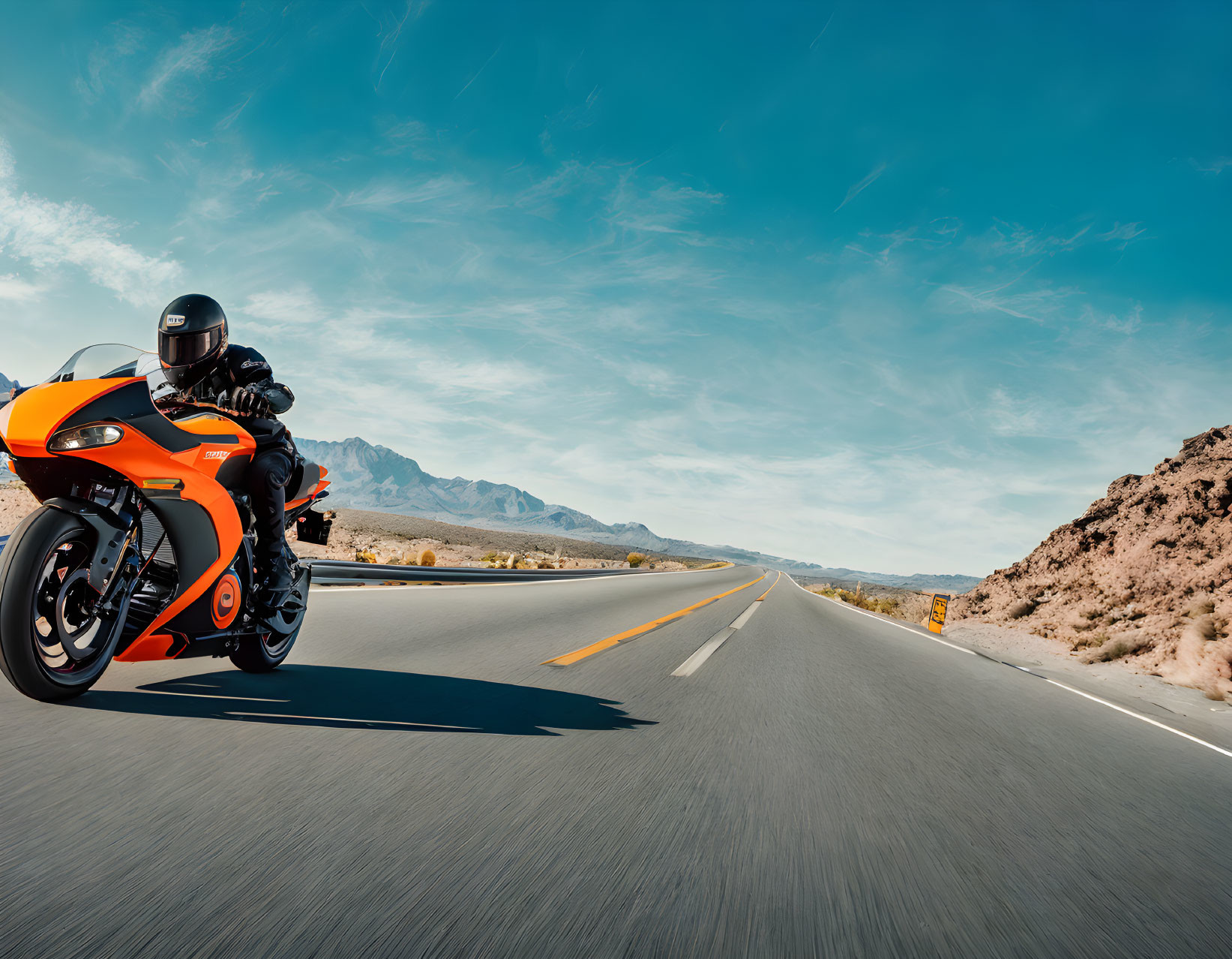 Black-gear motorcyclist on orange sportbike rides desert road under blue sky