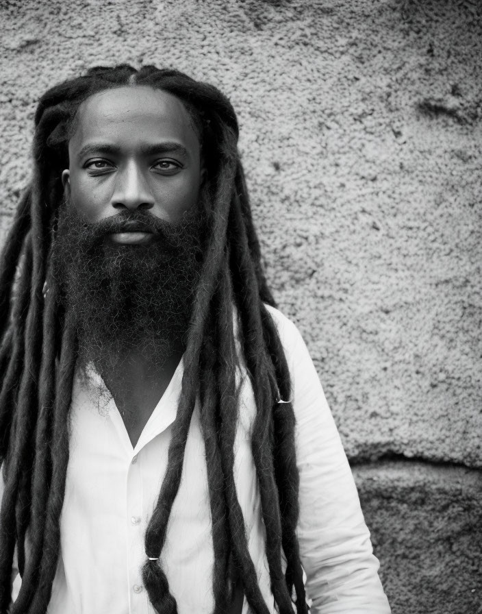 Man with long dreadlocks and beard in white shirt against textured wall