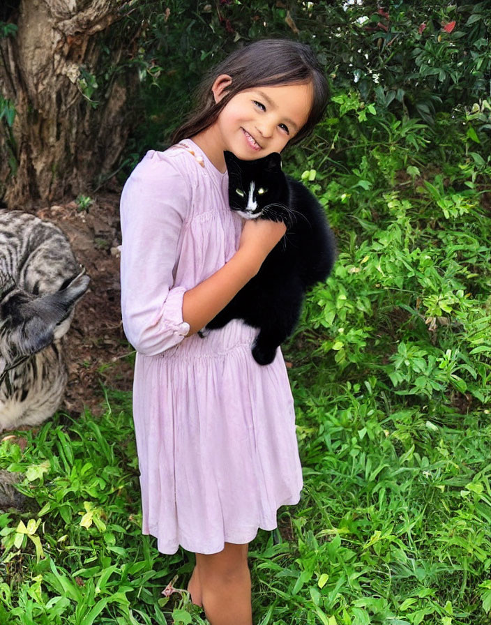 Young girl in pink dress holding black cat outdoors with green foliage and gray tabby cat.