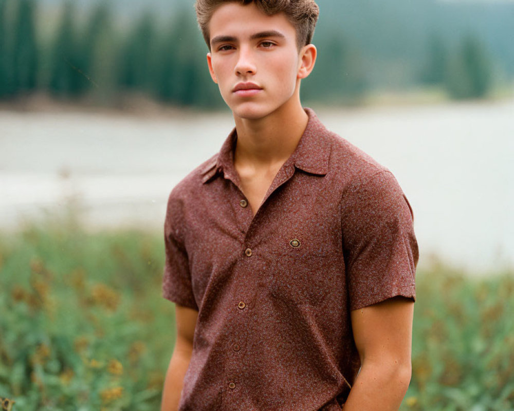 Young man in brown shirt posing by lake and mountains.