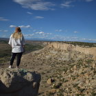 Person in hat and coat on rocky ledge overlooking vast flower-covered landscape under cloudy sky