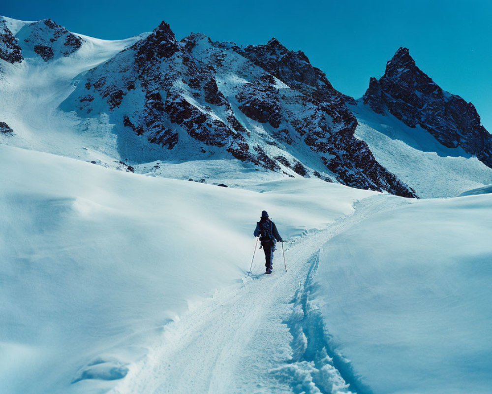 Snow-covered slope with lone skier and mountain peaks under clear blue sky