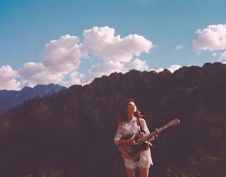 Person holding a guitar in sunlit mountainscape with fluffy clouds