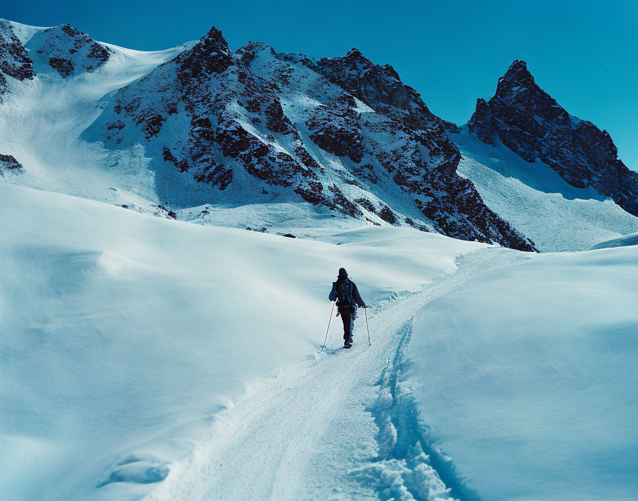 Snow-covered slope with lone skier and mountain peaks under clear blue sky