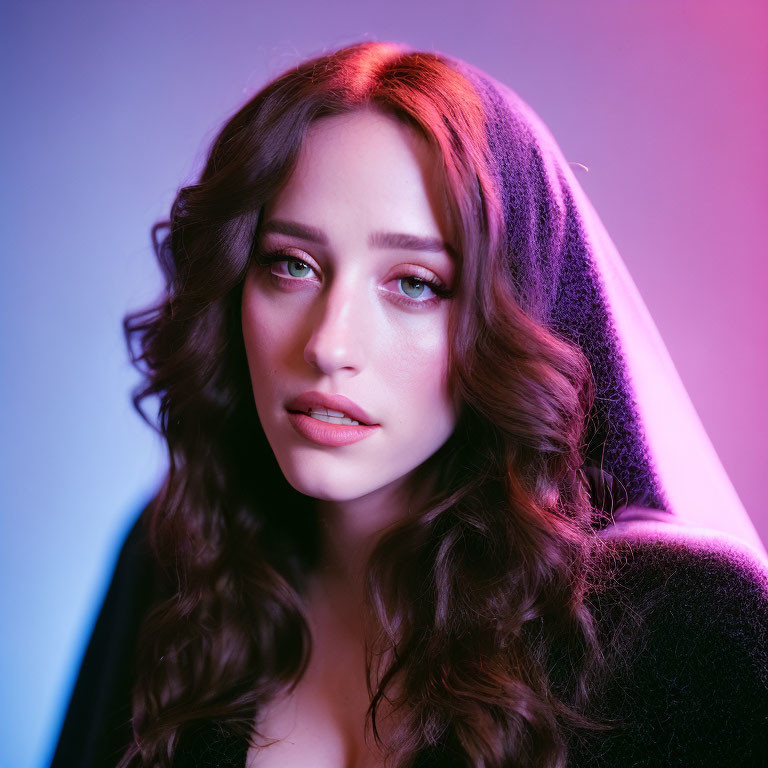 Young woman with blue eyes and dark wavy hair under pink and blue lighting gazes contemplatively.