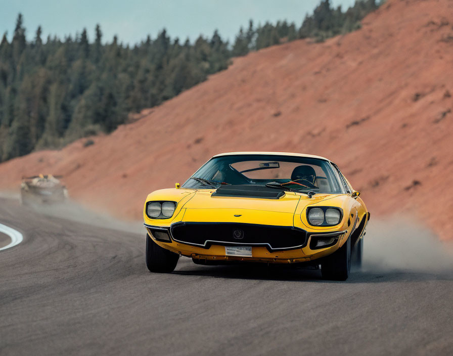 Yellow vintage sports car with black stripes racing on track with smoking tires, second car trailing in pine tree