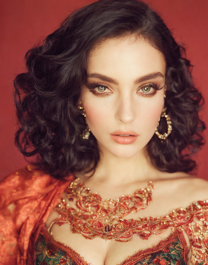 Woman with dark wavy hair, green eyes, gold jewelry, and embroidered dress on red background