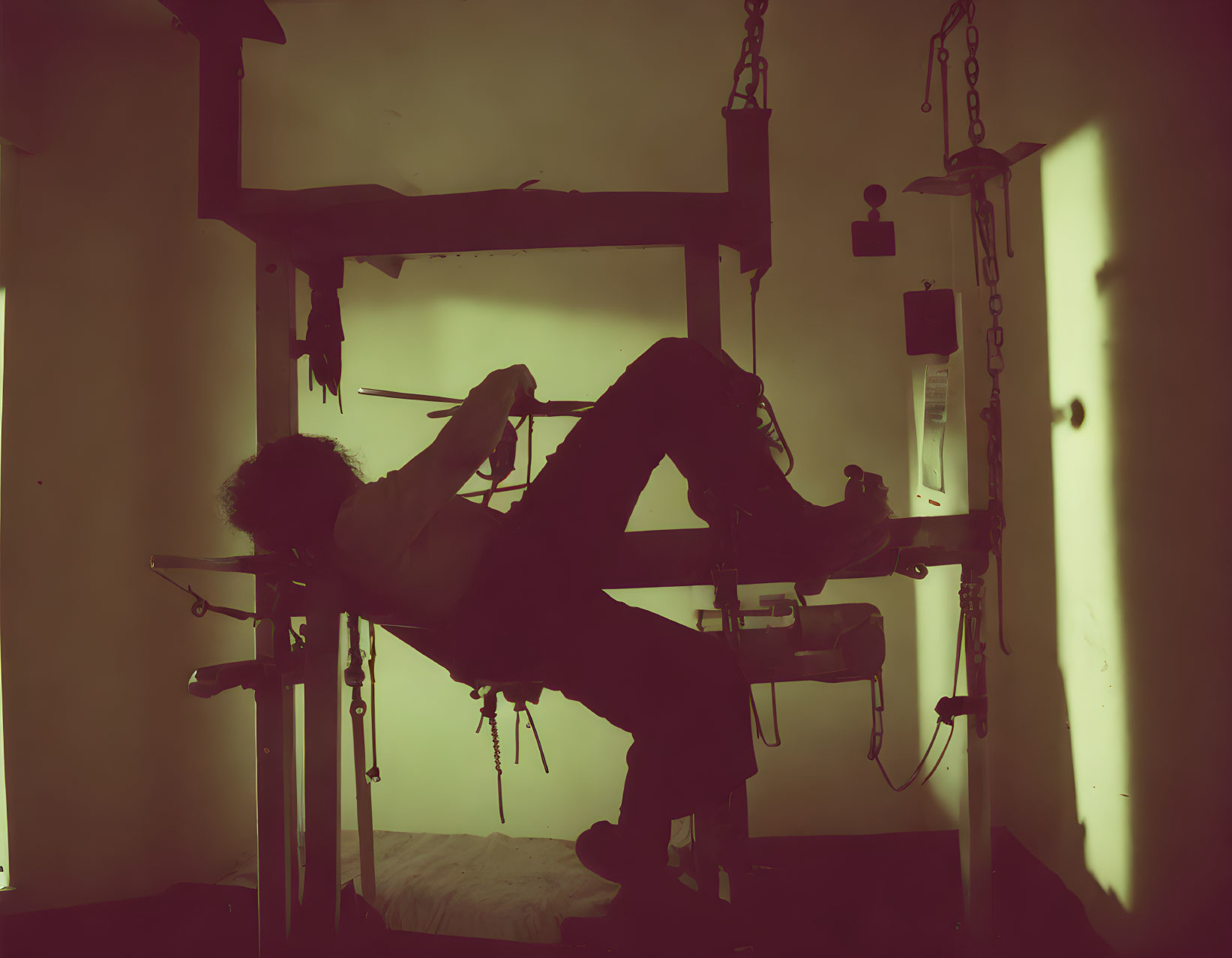 Person hanging upside down on chains in dim, yellow-lit room