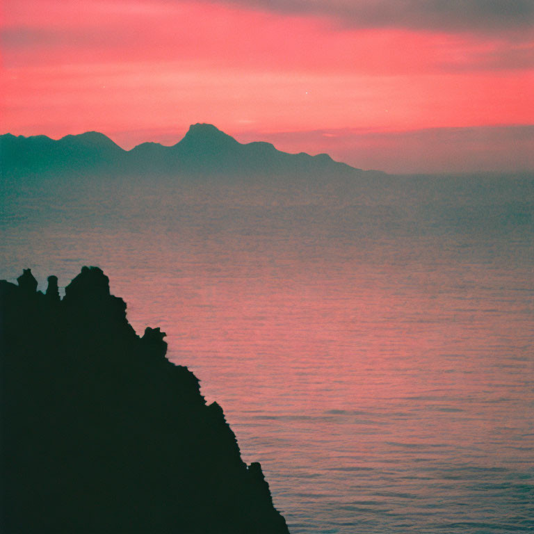 Colorful pink and red sunset over calm sea with silhouetted figures on cliff and distant mountains