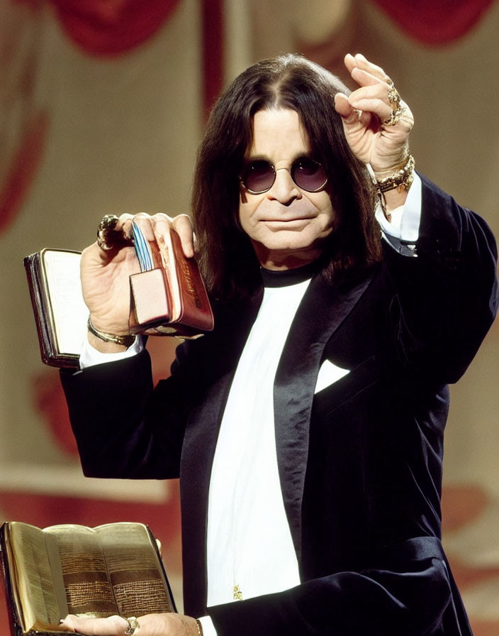 Dark-haired person in black suit with book and small item, standing by red curtain