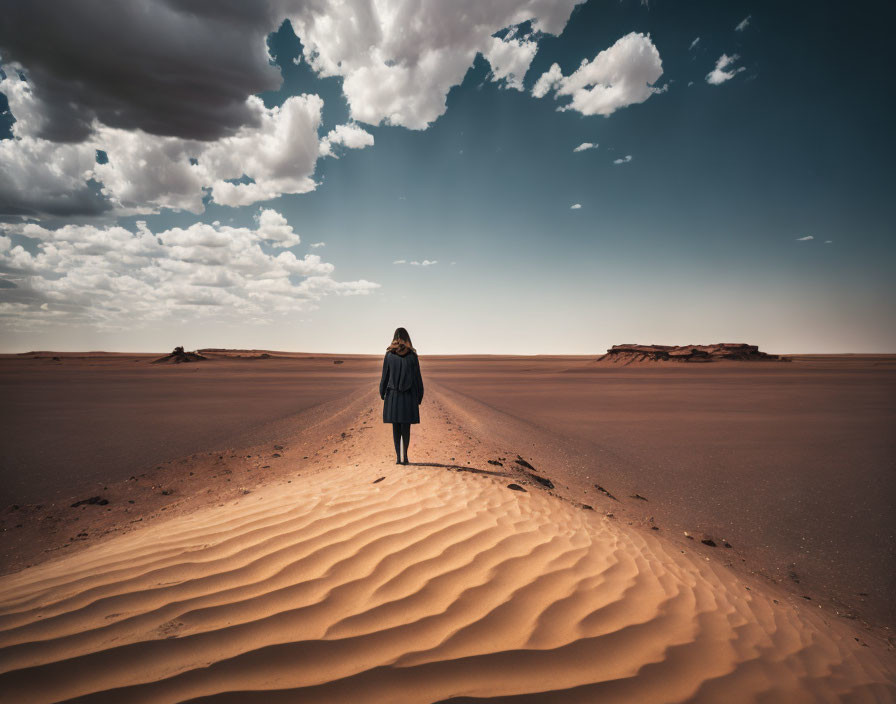 Person standing in desert sands under vast sky with scattered clouds