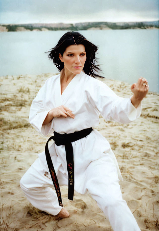 Person in white karate gi and black belt striking a pose on sandy beach with calm lake and over