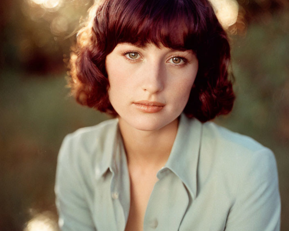 Woman with short curly brown hair in light blue shirt gazes at camera in serene setting