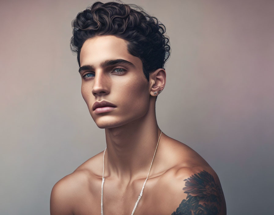 Young man with curly hair, blue eyes, earring, and tattoo on shoulder portrait.