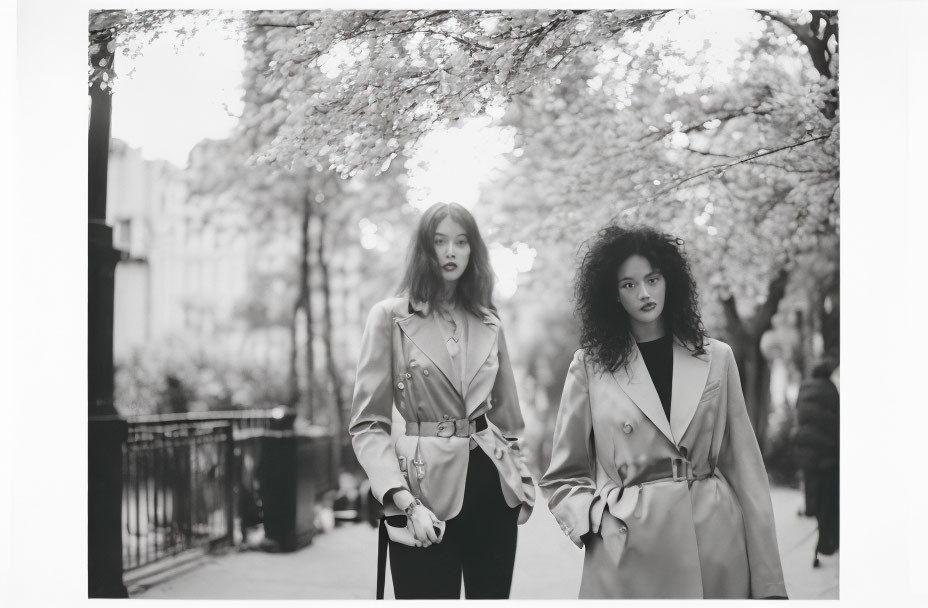 Stylish women in trench coats on tree-lined path with blooming branches