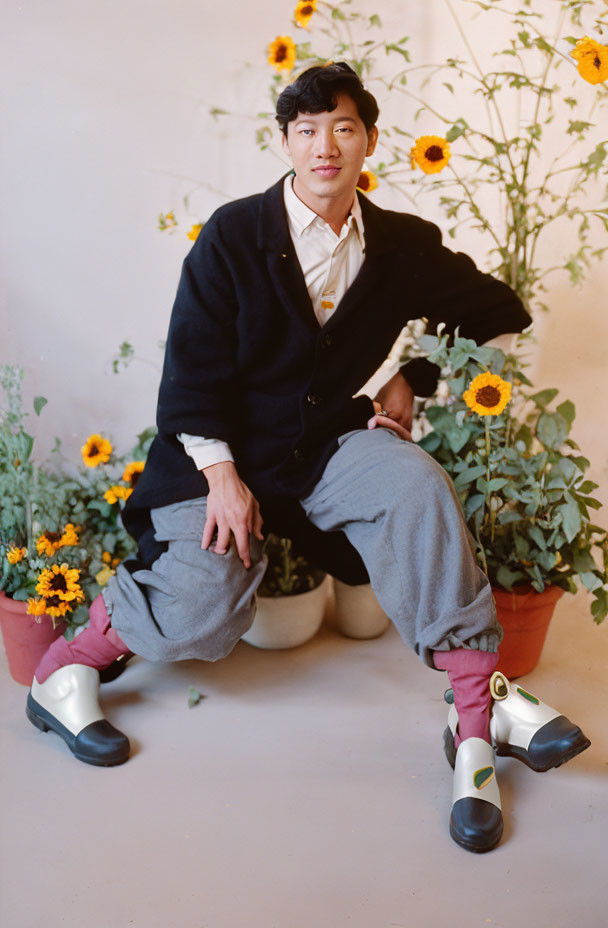 Seated person in navy jacket and grey pants with sunflower backdrop and white/green boots