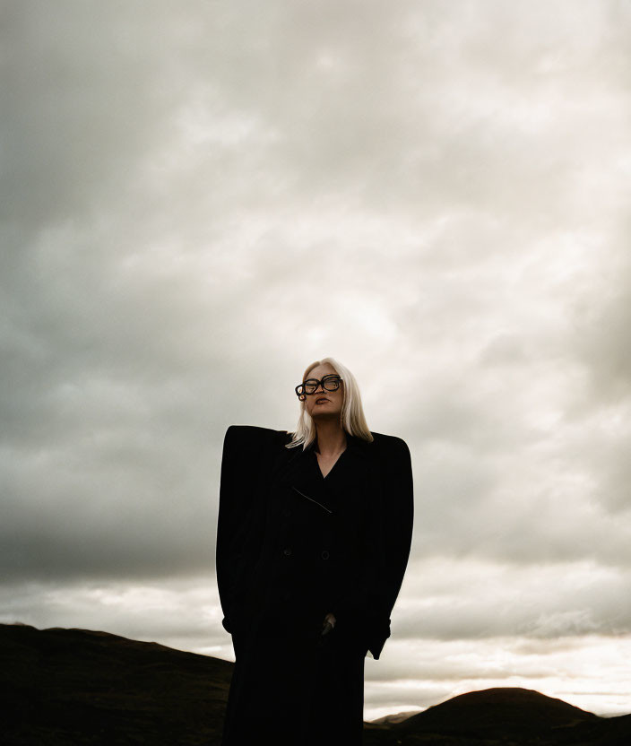 Person in Black Coat and Glasses Under Dramatic Cloudy Sky