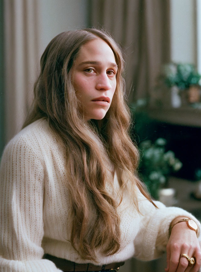 Young woman in white sweater with long wavy hair, posing in room with plants.