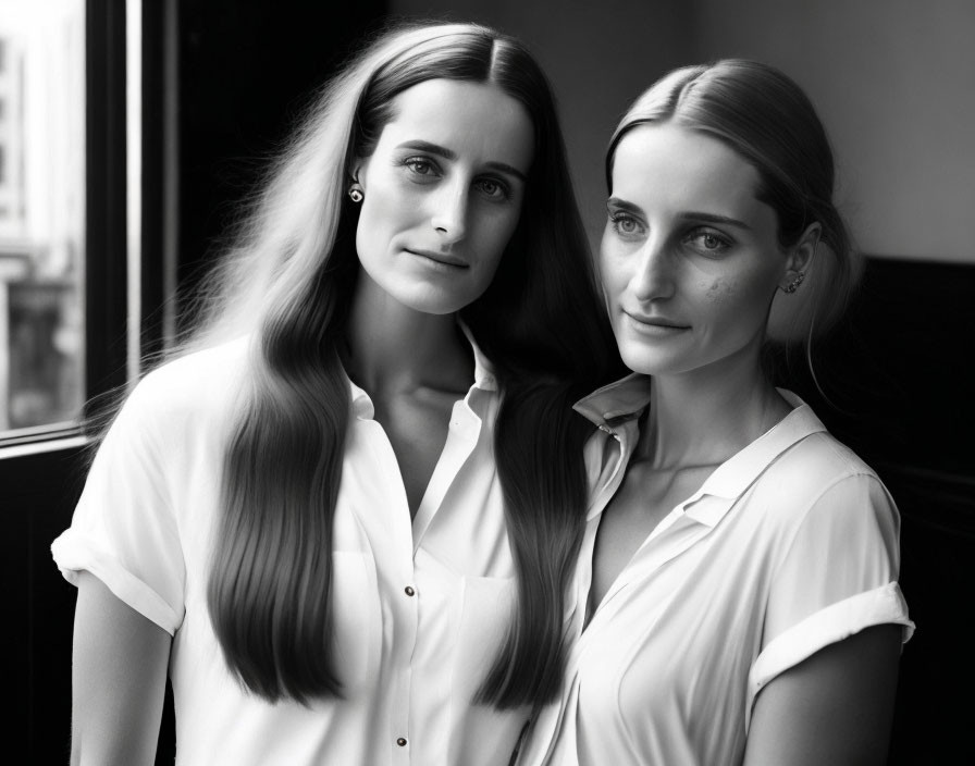 Two Women in White Shirts Pose by Window in Grayscale Photo