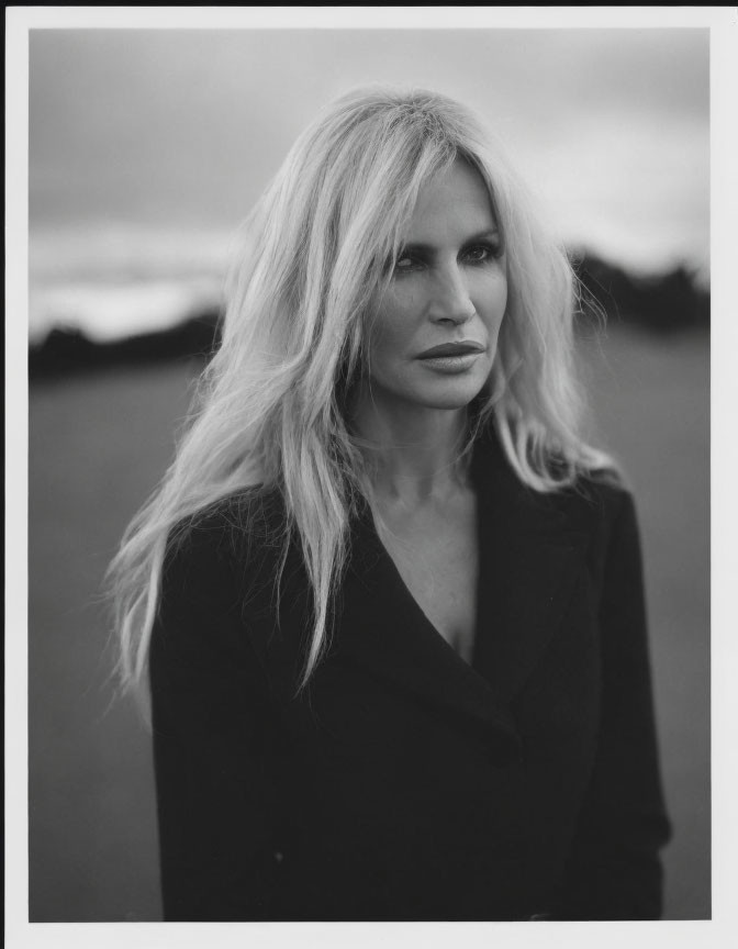 Monochrome portrait of woman with long blonde hair and dark blazer against cloudy sky.