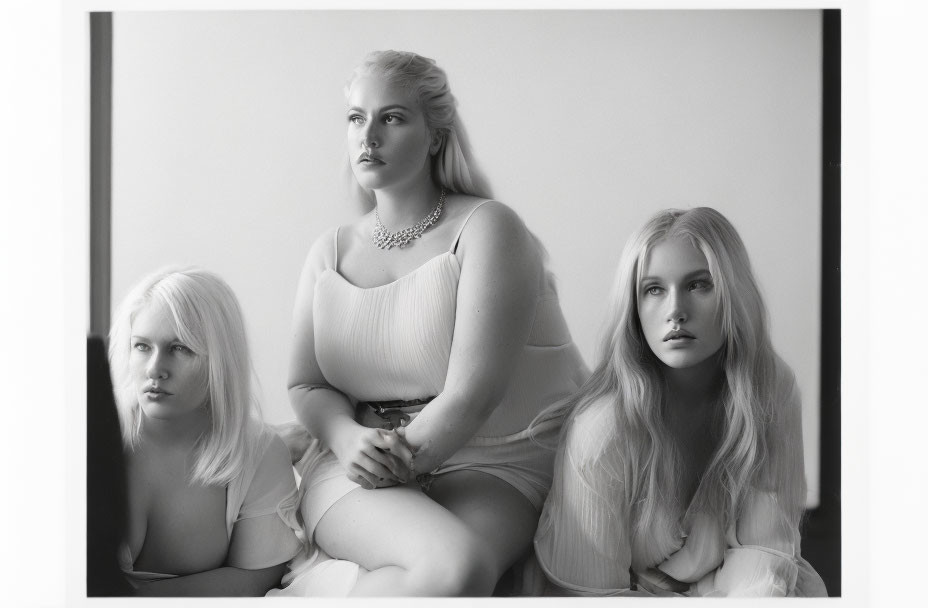 Three women with blonde hair in different styles, dressed in light clothing, sitting together solemnly.