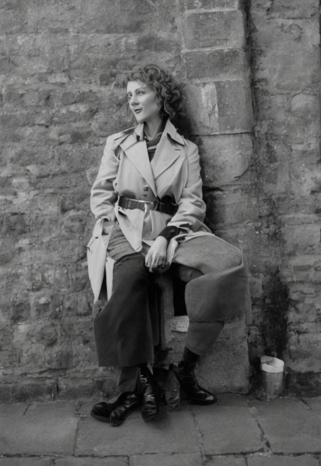 Woman in trench coat smiling on ledge against textured stone wall