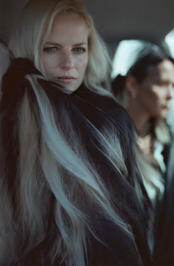 Blond woman with intense gaze in black attire, second person out of focus