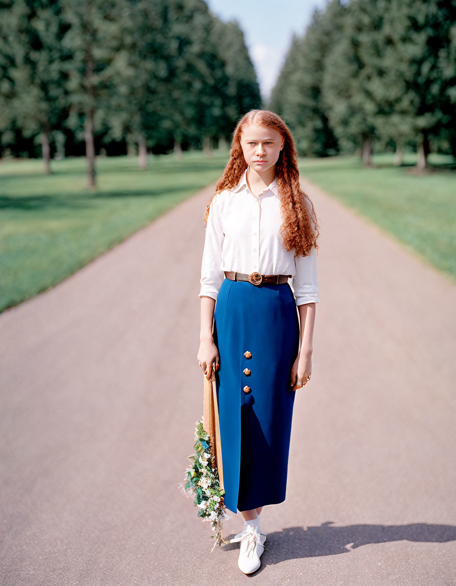 Young woman with long red hair holding flowers in park pathway outfit description.