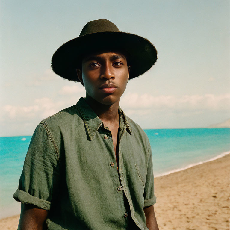 Neutral expression person in dark hat and green shirt on beach with clear sky.