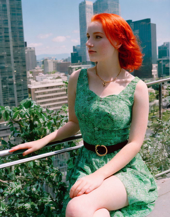 Red-haired woman in green dress sitting on railing with cityscape view.